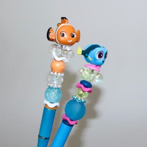 May include: Two blue pens with decorative tops featuring cartoon fish characters. One pen has an orange fish, the other a blue fish. The pens have beaded accents in various colors, including clear, orange, and blue, with a glittery blue bead.