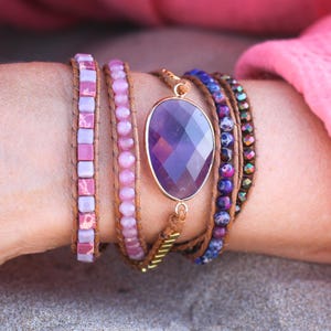 May include: A collection of wrap bracelets in shades of pink, purple, and brown. One bracelet features a large, oval amethyst gemstone pendant with a gold-colored setting. Other bracelets have pink beads and square tiles.