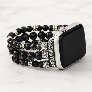 Peut inclure: Un bracelet de montre connectée composé de trois rangées de perles en pierre noire et gris foncé mouchetées. Les rangées sont reliées au boîtier par des éléments métalliques argentés aux motifs gravés et des perles intercalaires.