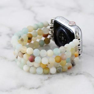 Puede incluir: Un reloj inteligente plateado con una pulsera de cuentas de varios hilos. La pulsera presenta cuentas redondas en tonos de blanco, amarillo, verde y marrón. El reloj es parcialmente visible, mostrando la pantalla y la fijación de la pulsera.