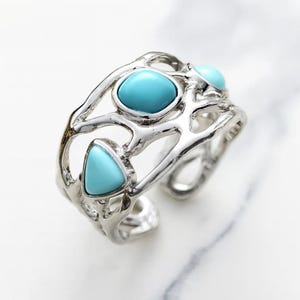 Puede incluir: Un brazalete plateado con un diseño calado único. El brazalete presenta tres piedras de color turquesa en formas ovaladas y triangulares, engastadas en la intrincada filigrana metálica. El brazalete está abierto en la parte inferior.