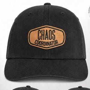 May include: A black baseball cap featuring a tan, shield-shaped patch on the front with the text "CHAOS COORDINATOR" in black lettering. The cap includes a curved brim, a six-panel crown, and an adjustable strap with a metal buckle at the back.