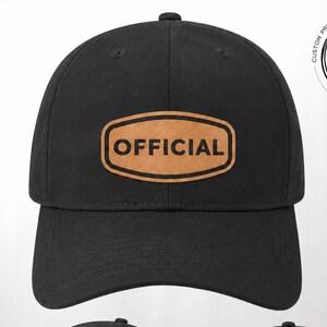 May include: A black baseball cap featuring a tan, oval-shaped patch on the front and side with the word OFFICIAL printed in black. The cap has a curved brim and an adjustable strap at the back. The text LEGACY BLACK - OFFICIAL PATCH is below.