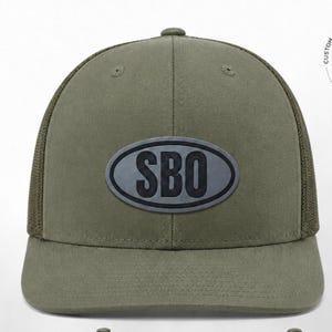 May include: An olive green baseball cap featuring a structured front panel and a mesh back. Centered on the front is an oval-shaped patch with a dark grey border and background, displaying the bold, black, capitalized letters SBO in the center.
