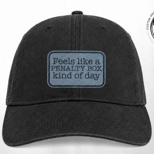 May include: A black baseball cap featuring a rectangular grey patch on the front. The patch contains the dark text "Feels like a PENALTY BOX kind of day" in a serif font. The cap has a structured crown, a curved brim, and visible stitching.