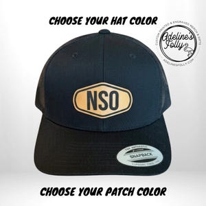 May include: A black trucker hat featuring a tan hexagonal patch with the letters NSO in black. The hat includes a flat brim with a circular sticker that reads The Authentic Yupoong Snapback. Text above reads CHOOSE YOUR HAT COLOR.