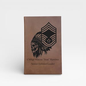 May include: A brown leatherette notebook with a black graphic of a skull wearing a Native American headdress and military insignia. The text reads "CMSgt Marcus "Iron" Ramirez Senior Enlisted Leader."