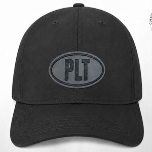 May include: A solid black baseball cap featuring a centered oval patch with the letters PLT embossed in a bold, dark font. The cap has a structured crown, a curved brim with visible stitching, and a classic six-panel design with top eyelets.