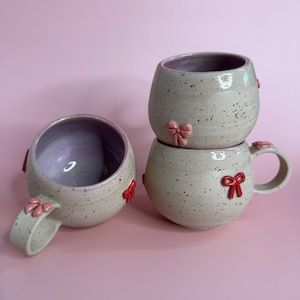 Könnte beinhalten: Drei gesprenkelte Keramikbecher mit rosa Innenräumen und roten Schleifenakzenten. Eine Tasse liegt auf der Seite und zeigt das Innere. Die anderen beiden Tassen sind gestapelt. Die Tassen haben eine abgerundete Form und einen Henkel.