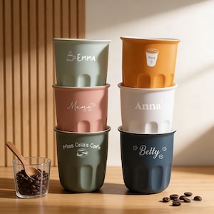 Könnte beinhalten: Ein Stapel von sechs bunten Keramik-Kaffeetassen. Jede Tasse hat eine andere Farbe, darunter Grün, Orange, Rosa, Weiß und Dunkelblau. Auf jeder Tasse ist ein Name in Weiß aufgedruckt, wie z. B. "Emma" und "Anna". Ein kleines Glas mit Kaffeebohnen und ein Holzlöffel befinden sich neben den Tassen.