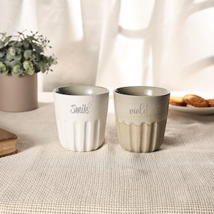 May include: Two ceramic mugs, one white and one beige, sit on a textured surface. The white mug has the word "Smile" and a star design, while the beige mug has the word "violet". Both mugs have a ribbed design.