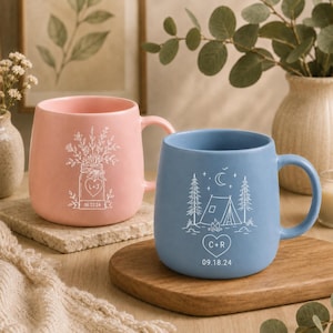 Puede incluir: Una taza de cerámica rosa claro con un dibujo lineal blanco de un ramo de flores en un frasco con el texto L+J y 06.22.24, y una taza azul claro con un dibujo lineal blanco de una tienda de campaña, árboles y luna con C+R y 09.18.24.