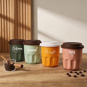 May include: Four brightly coloured travel mugs with dark brown lids and white text. The mugs are green, light green, orange, and pink. The text on the mugs reads "Emma", "Anna", "Mama", and "PAPA". A small glass cup with coffee beans and a wooden spoon is in the foreground.