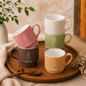 May include: A set of five ceramic mugs in various matte colors including dusty rose, dark brown, cream, sage green, and mustard yellow. Each mug features a white line-art floral illustration and a name: Olivia, Amelia, Emma, Lily, and Sophia.