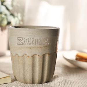 Könnte beinhalten: Eine beige Keramiktasse mit dem Aufdruck "ZANNA" auf der Vorderseite. Die Tasse hat eine geriffelte untere Hälfte und eine glatte obere Hälfte. Die Tasse steht auf einer strukturierten Oberfläche.