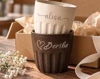 Puede incluir: Dos tazas de cerámica con texturas acanaladas verticales. La taza superior es de color crema con el nombre alisa grabado en cursiva. La taza inferior es de color marrón oscuro con un icono de corazón y el nombre Bertha grabado.