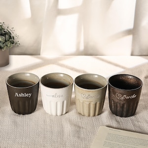 May include: Four ceramic mugs with personalized names: "Ashley", "alisa", "Bertha", and "Bertha". The mugs are in shades of brown, white, and beige, with a ribbed design. The mugs are on a neutral-colored surface.