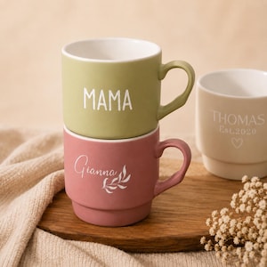 Puede incluir: Tazas de cerámica apiladas en verde oliva y rosa empolvado. La taza superior dice "MAMA" en blanco, mientras que la taza inferior dice "Gianna" con un diseño de hojas. También se ve una tercera taza beige.