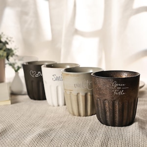 May include: A collection of five ceramic mugs in varying colors, including brown, white, and beige. Each mug features a unique inscription, such as "Smile" and "Grace and Faith". The mugs have a ribbed design and are arranged on a neutral-toned surface.