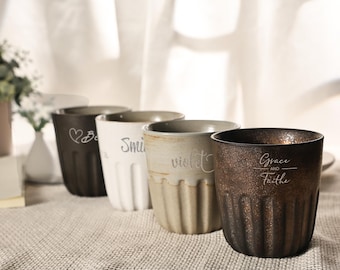 Puede incluir: Cuatro tazas de cerámica con texturas acanaladas verticales dispuestas en fila. Las tazas presentan diferentes colores e inscripciones: una taza marrón oscuro con un corazón, una blanca con la palabra Smile, una beige con violet y una bronce oscuro con Grace and Faithe.