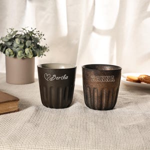 May include: Two dark brown, textured cups with the names "Bertha" and "Zanna" etched on them. The cup on the left has a heart symbol before the name. The cups are on a textured, off-white surface.