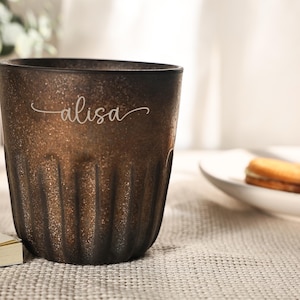 May include: A brown textured glass with vertical grooves. The word "alisa" is written in a cursive font on the glass. A white plate with a cookie is in the background.