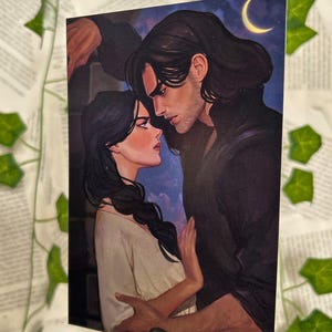 以下が含まれることがあります： 男性と女性がお互いを見つめ合っているロマンチックなイラストのプリント。男性は長い黒髪で、暗いコートを着ています。女性は長い黒髪で、明るい色のトップスを着ています。背景には三日月があります。