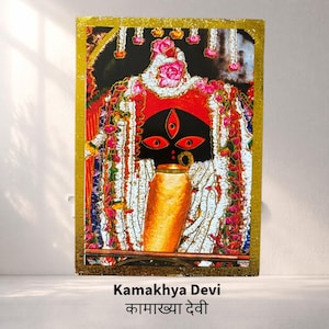 Puede incluir: Una impresión enmarcada de Kamakhya Devi, que presenta una deidad con rostro negro, ojos rojos y un recipiente dorado. La imagen está adornada con guirnaldas florales y enmarcada en oro, con texto en la parte inferior.