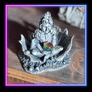 May include: A silver-colored fairy figurine seated on a leaf-like base, holding a colorful crystal ball. The fairy has detailed features and is surrounded by intricate leaf designs. The figurine is displayed on a wooden surface.
