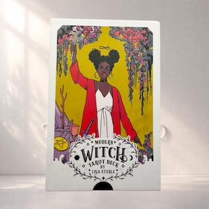 May include: A tarot deck box with an illustration of a Black woman in a red jacket and white dress, holding a wand. The box has the text "Modern Witch Tarot Deck by Lisa Sterle" with floral accents and a yellow background.