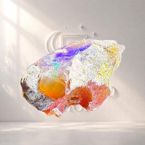 Op de afbeelding: Een kleurrijke, abstracte edelsteen met een ruw, getextureerd oppervlak. De steen vertoont een spectrum aan kleuren, waaronder oranje, rood, paars en geel, met een witte, kristallijne buitenkant. De afbeelding is geplaatst tegen een witte achtergrond met een maan- en sterrenontwerp.