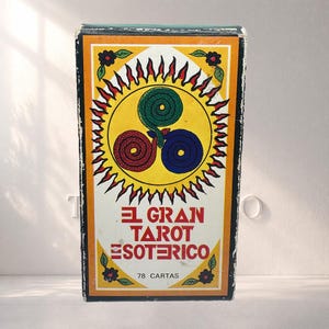 Pode incluir: Uma caixa de cartas de tarô vintage com um design amarelo, laranja e preto. A caixa apresenta o texto "EL GRAN TAROT ESOTERICO" e uma ilustração de três cobras entrelaçadas em vermelho, verde e azul. A caixa também diz "78 CARTAS".
