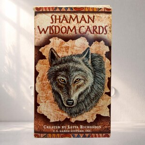 May include: A deck of "Shaman Wisdom Cards" featuring a detailed illustration of a wolf's head with amber eyes. The card box has a tan background with a torn paper effect. The text "SHAMAN WISDOM CARDS" is at the top.