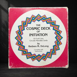 May include: A square box for The Cosmic Deck of Initiation, featuring a white centre with a rainbow-coloured geometric border. The text reads: The Cosmic Deck of Initiation, 52 Full-Colour Circular Mandala Cards by Barbara M. DeLong, Complete with Instructions. The background is a solid magenta pink.