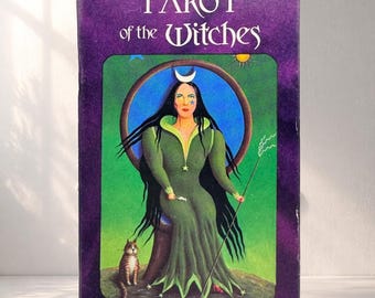 May include: A tarot card box with the title "TAROT of the Witches" in white lettering on a purple background. The box features an illustration of a woman in a green dress, a cat, and a staff.