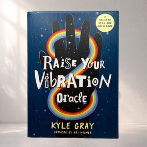 May include: A navy blue oracle deck box featuring the words "Raise Your Vibration Oracle" in white. A black hand with a rainbow arch is in the center. The text "A 48-Card Deck and Guidebook" is in the upper right corner. The author's name is "Kyle Gray".