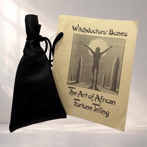 May include: A black drawstring pouch with a textured surface paired with a rectangular beige card. The card features the text Witchdoctors' Bones and The Art of African Fortune Telling above an illustration of a figure standing before stones.