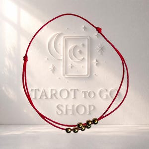 May include: A red string bracelet with gold beads. The bracelet is in front of a white background with the words "TAROT TO GO SHOP" embossed. A crescent moon and stars are also embossed on the background.
