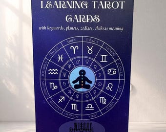 Puede incluir: Una caja azul marino de "Learning Tarot Cards" con palabras clave, planetas, signos del zodiaco y significados de los chakras. La caja presenta un diseño circular con símbolos del zodiaco y una figura central. El texto "Witchy Cauldron" está en la parte inferior.