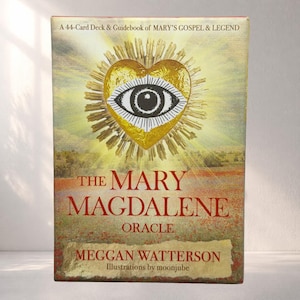 May include: A 44-card deck and guidebook titled The Mary Magdalene Oracle by Meggan Watterson with illustrations by moonjube. The box features a golden heart with an eye at the centre, set against a field of red flowers under a bright sky.