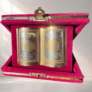 May include: A decorative, open book with gold and silver metal detailing, displayed inside a red velvet box. The book's pages feature intricate patterns and Arabic script. The box has a gold clasp and hinges.