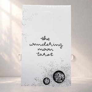 Puede incluir: Una caja rectangular blanca para cartas de tarot con el texto negro 'the wandering moon tarot' centrado en la parte frontal. El diseño incluye patrones de estrellas punteadas en negro en la esquina superior izquierda y dos ilustraciones circulares de lunas en la parte inferior.