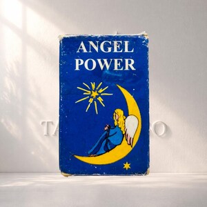 May include: A rectangular book titled ANGEL POWER with a deep blue cover. The illustration features a yellow crescent moon, a bright yellow star, and a figure with long blonde hair and white wings sitting on the moon in a blue robe.