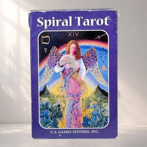 May include: A rectangular purple box for the Spiral Tarot deck. The front features a winged figure pouring liquid from a vessel into another, set against a rainbow sky, mountains, and blue flowers. The text reads Spiral Tarot, XIV, and U.S. Games Systems, Inc.