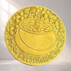 May include: A gold coin with a design of a pot filled with coins, encircled by shamrocks. The coin has the text "НА ЩАСТЯ І БЕЗПЕКУ!" around the edge. The design is embossed, giving it a three-dimensional appearance.