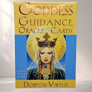 Pode incluir: Caixa retangular para "Goddess Guidance Oracle Cards" de Doreen Virtue. A caixa apresenta um retrato de uma mulher com coroa e colar de pedras azuis, sobre um fundo azul e dourado. O texto "A 44-Card Deck with Guidebook" também é visível.