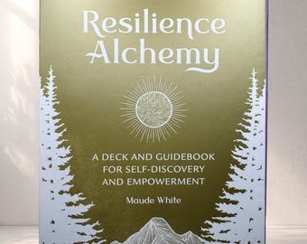 Op de afbeelding: Een boek getiteld "Resilience Alchemy" met een goud-witte omslag. De omslag toont de titel in witte letters, een zonnesterontwerp en de tekst "A Deck and Guidebook for Self-Discovery and Empowerment" en de naam van de auteur, Maude White.