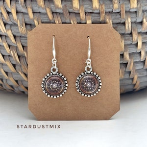 May include: A pair of silver-toned drop earrings featuring circular pendants with a dark brown and white mandala pattern. The pendants are framed by a beaded edge and hang from silver-toned hooks. The text STARDUSTMIX is printed below.