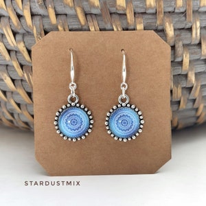May include: A pair of silver-toned drop earrings featuring circular charms with a blue and white mandala-style pattern. The charms are bordered by small silver beads and hang from silver hooks. The text STARDUSTMIX is printed below the earrings.