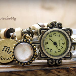 Puede incluir: Un reloj de cuero blanco con una esfera de color bronce y un dise&ntilde;o decorativo. El reloj tiene una esfera blanca con n&uacute;meros y manecillas negras. El reloj est&aacute; adornado con un brazalete de charms con una cuenta blanca, un charm de bronce con el s&iacute;mbolo astrol&oacute;gico de Virgo y la palabra "apasionado", y un peque&ntilde;o charm redondo blanco.